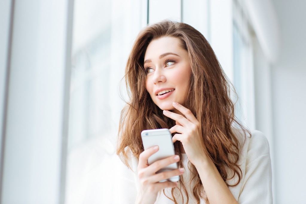 Portrait of a cheerful woman holding smartphone and looking at window